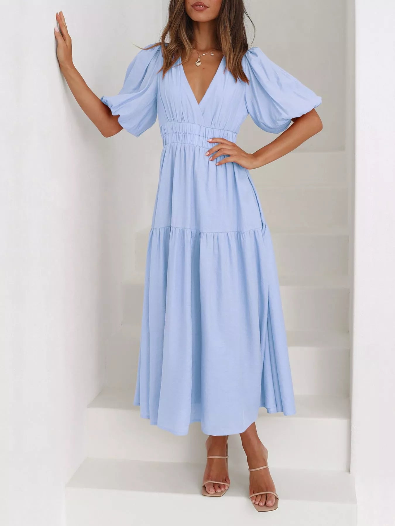 A woman wearing a light blue midi dress with puff sleeves, a V-neckline, and a tiered skirt, standing on white stairs.