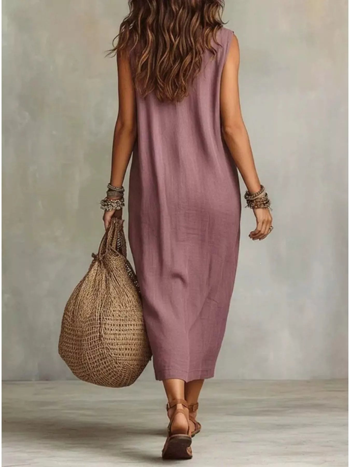 A woman shown from behind wearing a sleeveless, midi-length dusty mauve dress and carrying a large woven straw tote bag against a textured gray studio background.