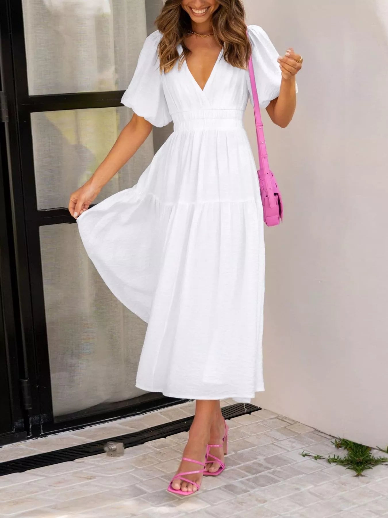 A woman wearing a white midi dress with puff sleeves and a V-neckline, carrying a bright pink shoulder bag and wearing pink strappy heels, standing in front of a light-colored wall and glass door.