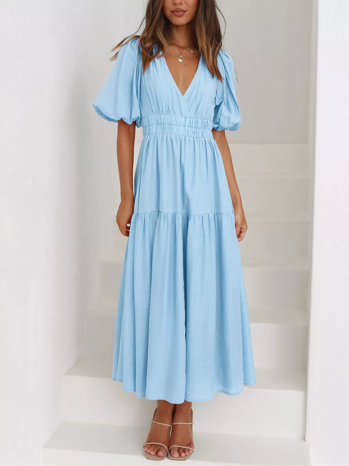 A woman wearing a light blue midi dress with puff sleeves, a V-neckline, and a smocked waist, standing on white stairs.