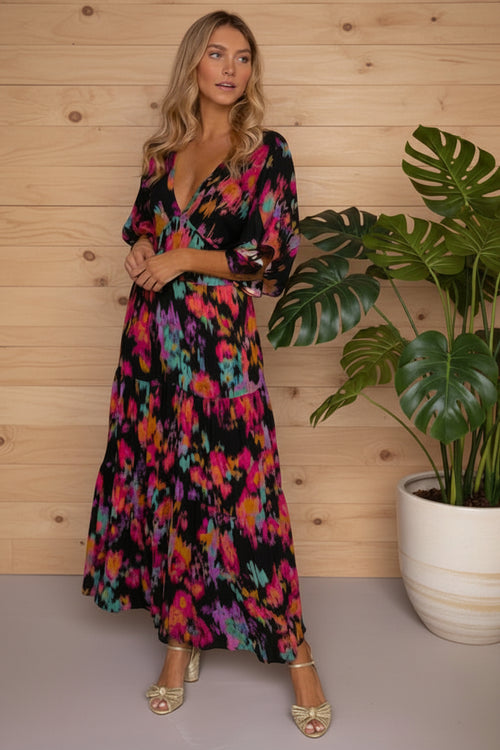 Woman wearing a colorful dress standing next to a potted plant against a wooden wall.