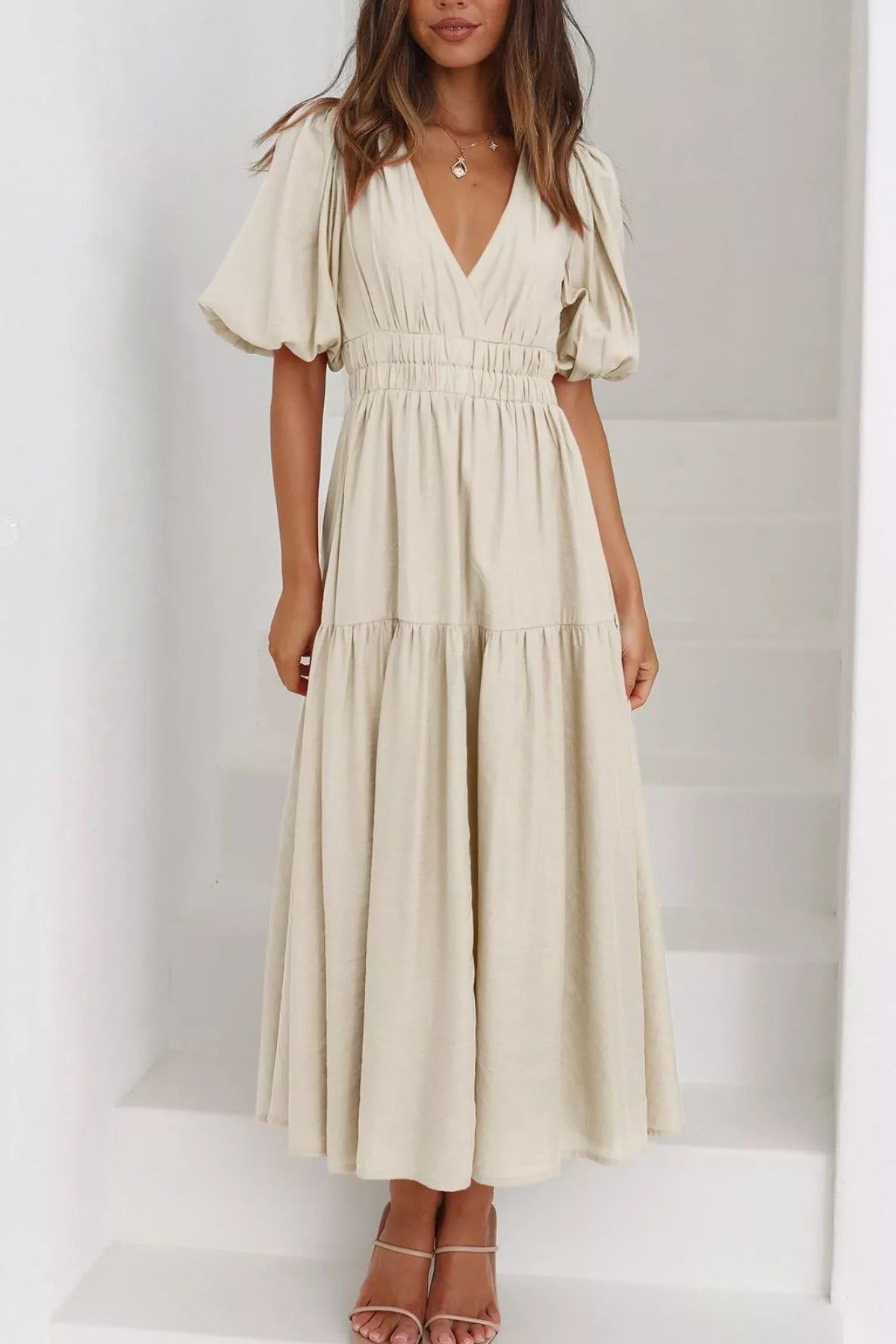 A woman wearing a light beige tiered maxi dress with puff sleeves and a V-neckline, standing on white stairs.
