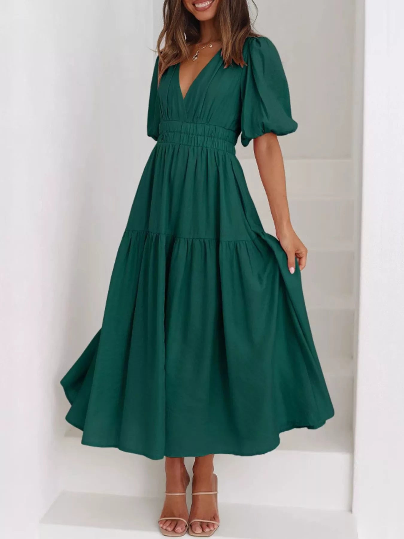 A woman wearing a dark green midi dress with puff sleeves, a deep V-neckline, and a tiered skirt, standing on white stairs.