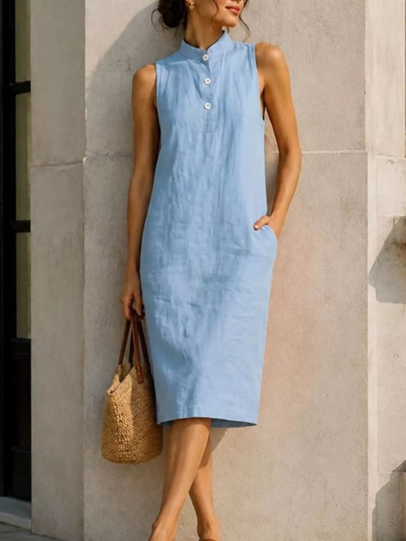 A woman wearing a sleeveless light blue midi dress with a high collar and button placket, standing against a light gray stone wall and holding a woven straw tote bag.