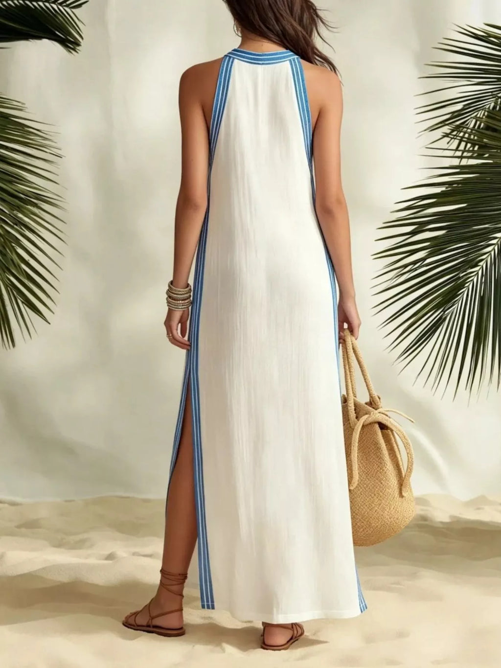 A woman stands on a sandy surface wearing a white sleeveless maxi dress with light blue trim and side slits, holding a woven straw bag. Palm leaves frame the scene against a light neutral backdrop.