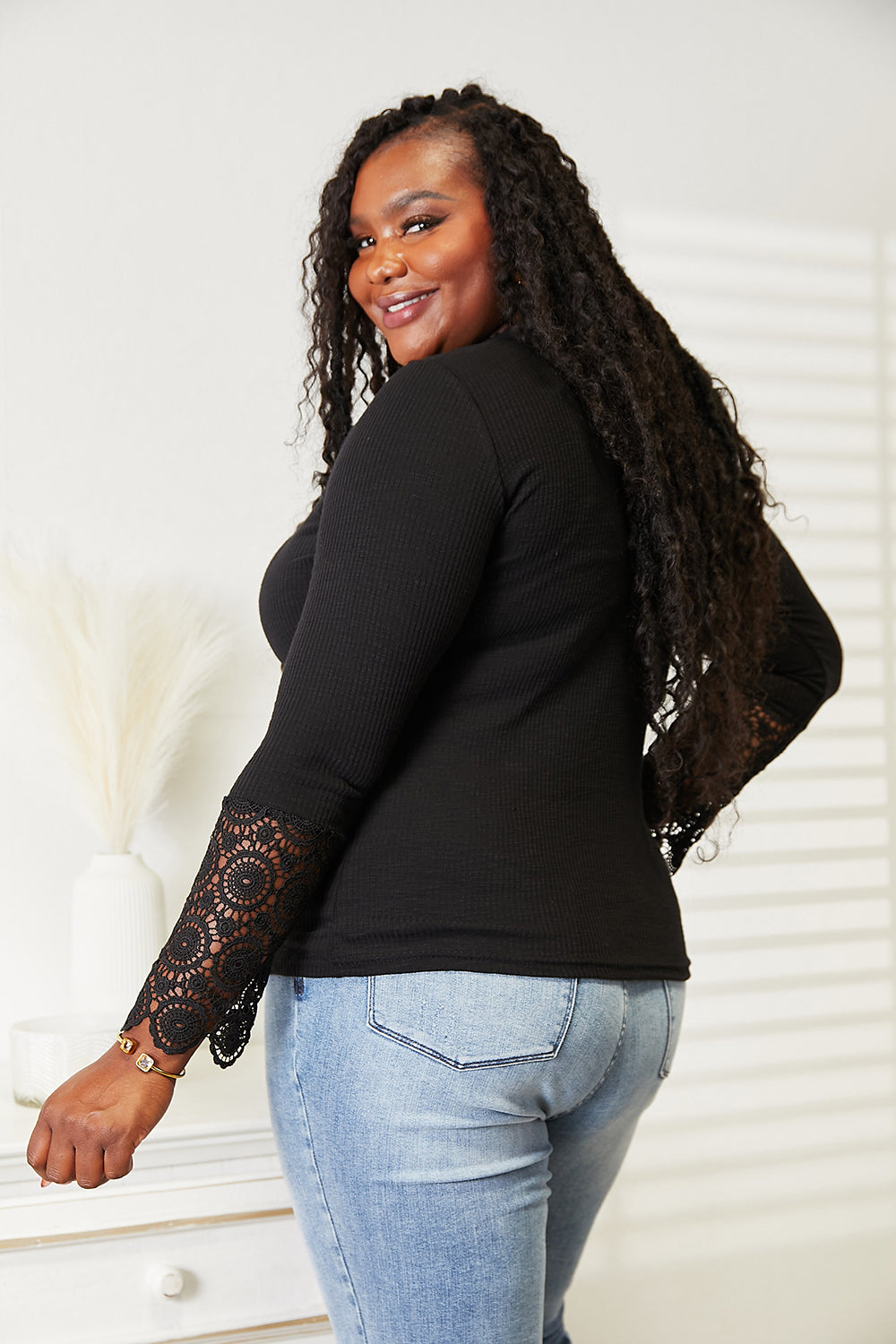 Woman wearing a black long-sleeve top with lace cuffs and light blue jeans against a white wall.