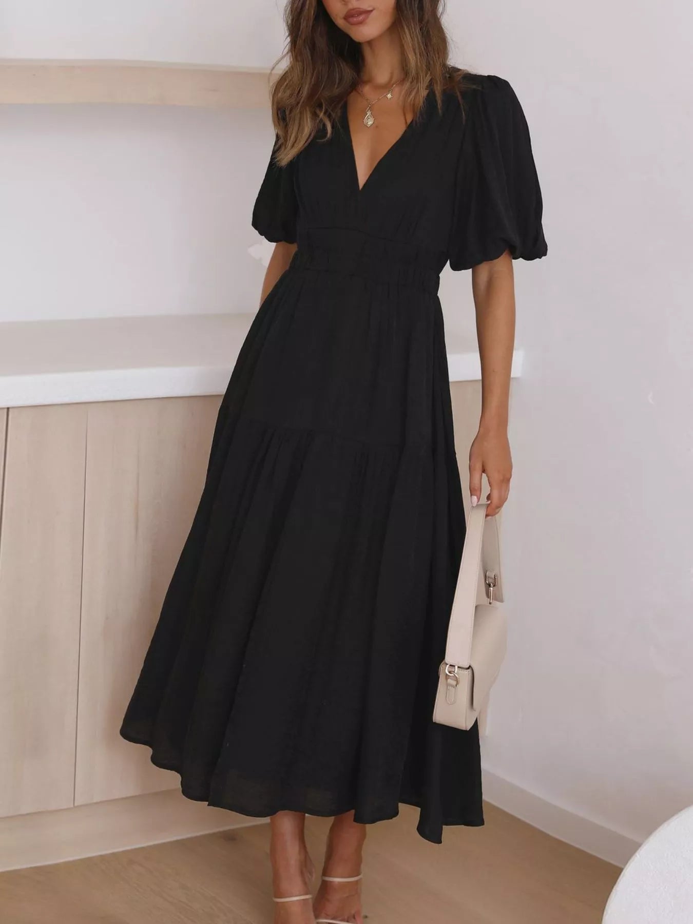 A woman wearing a black midi dress with puff sleeves, a deep V-neckline, and a tiered skirt, holding a cream-colored handbag in a bright indoor setting.