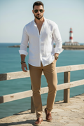 A man wearing a white linen button-down shirt, tan trousers, brown leather loafers, and sunglasses stands on a stone walkway next to a wooden railing with the ocean in the background.