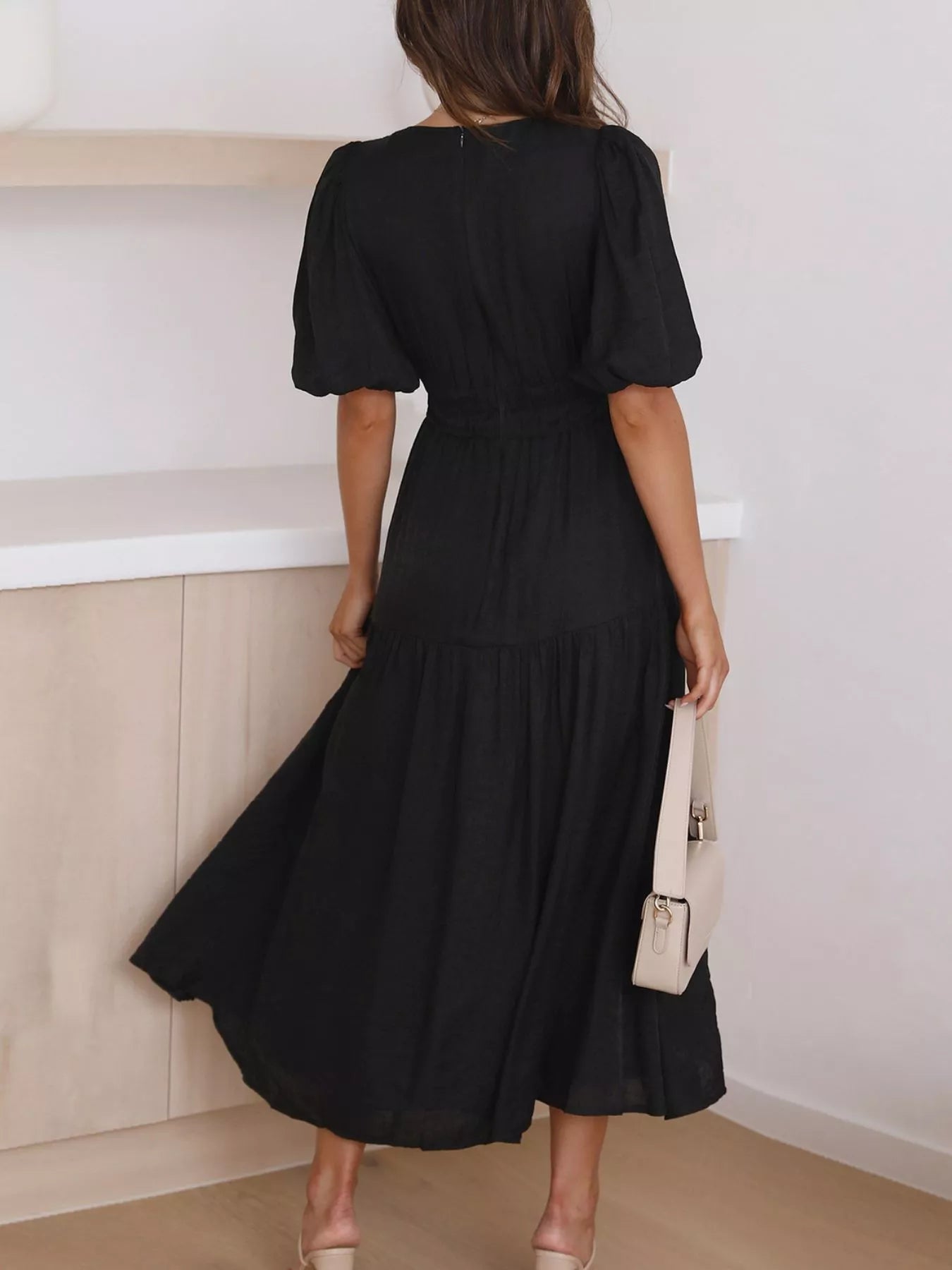 A woman wearing a black midi dress with puff sleeves and a tiered skirt, shown from the back in an indoor setting.