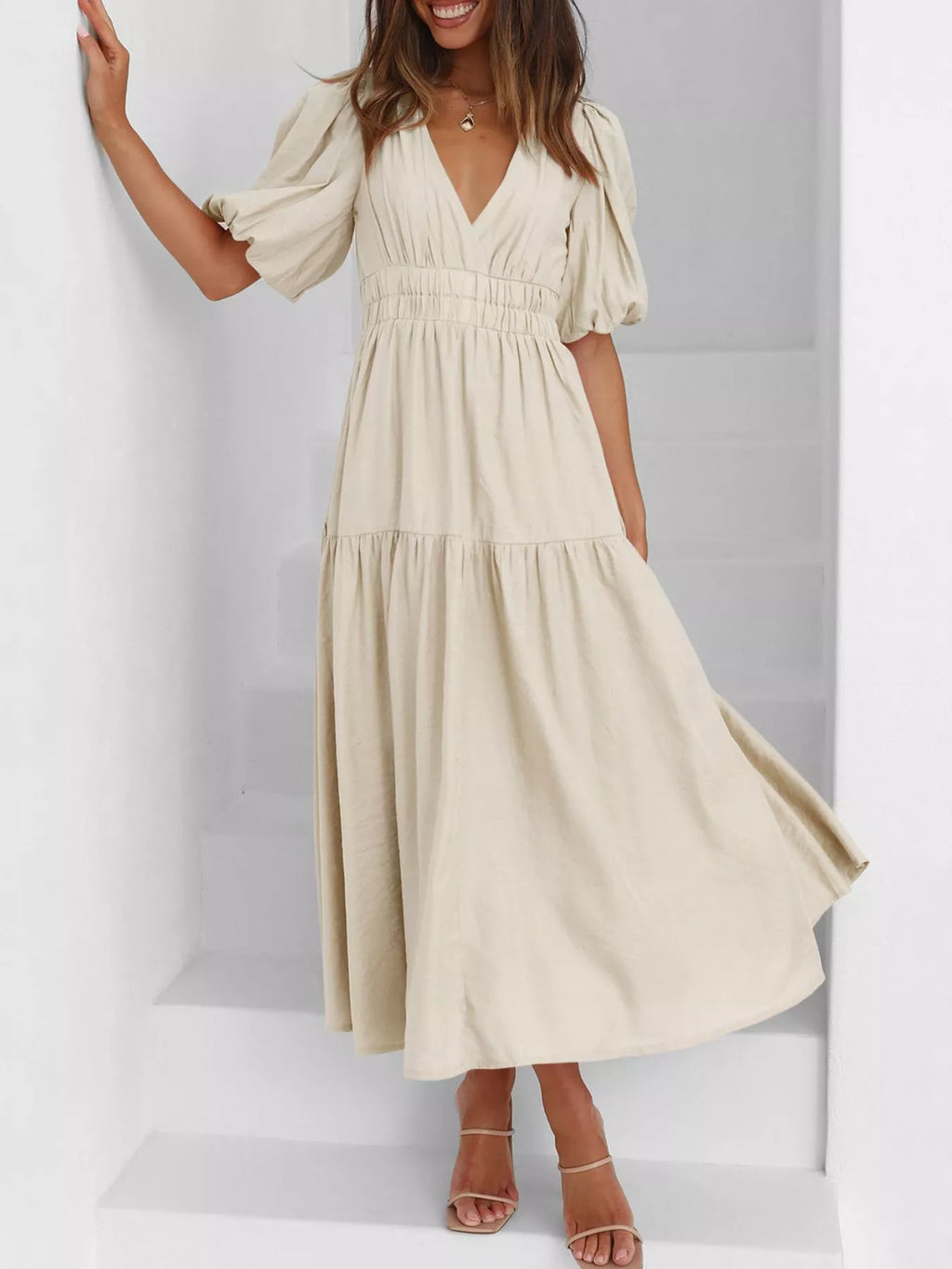 A woman wearing a cream-colored midi dress with puff sleeves, a V-neckline, and a tiered skirt, standing on white stairs.