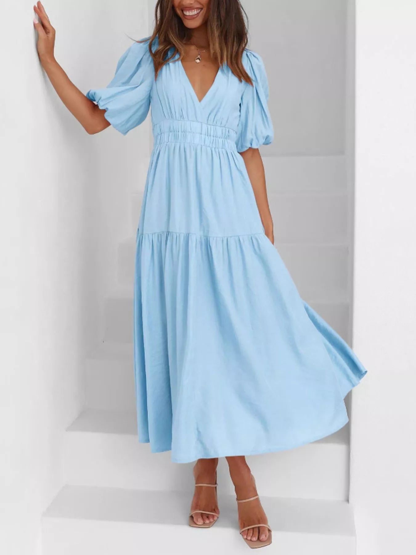 A woman wearing a light blue midi dress with puff sleeves, a V-neckline, and a tiered skirt, standing on white stairs.