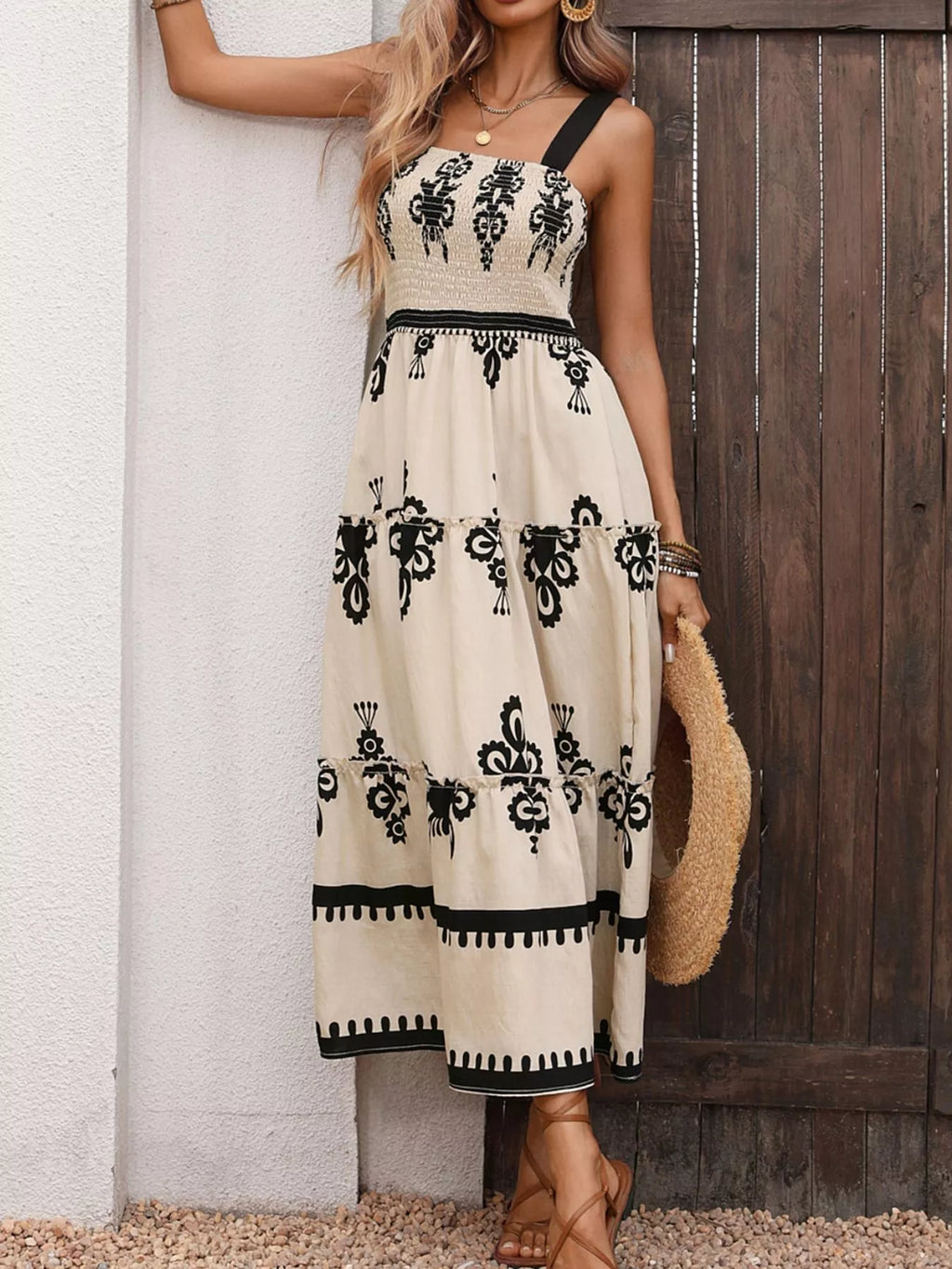 A woman models a sleeveless, one-shoulder maxi dress with a cream base and black floral patterns, standing against a white wall and dark wooden door.