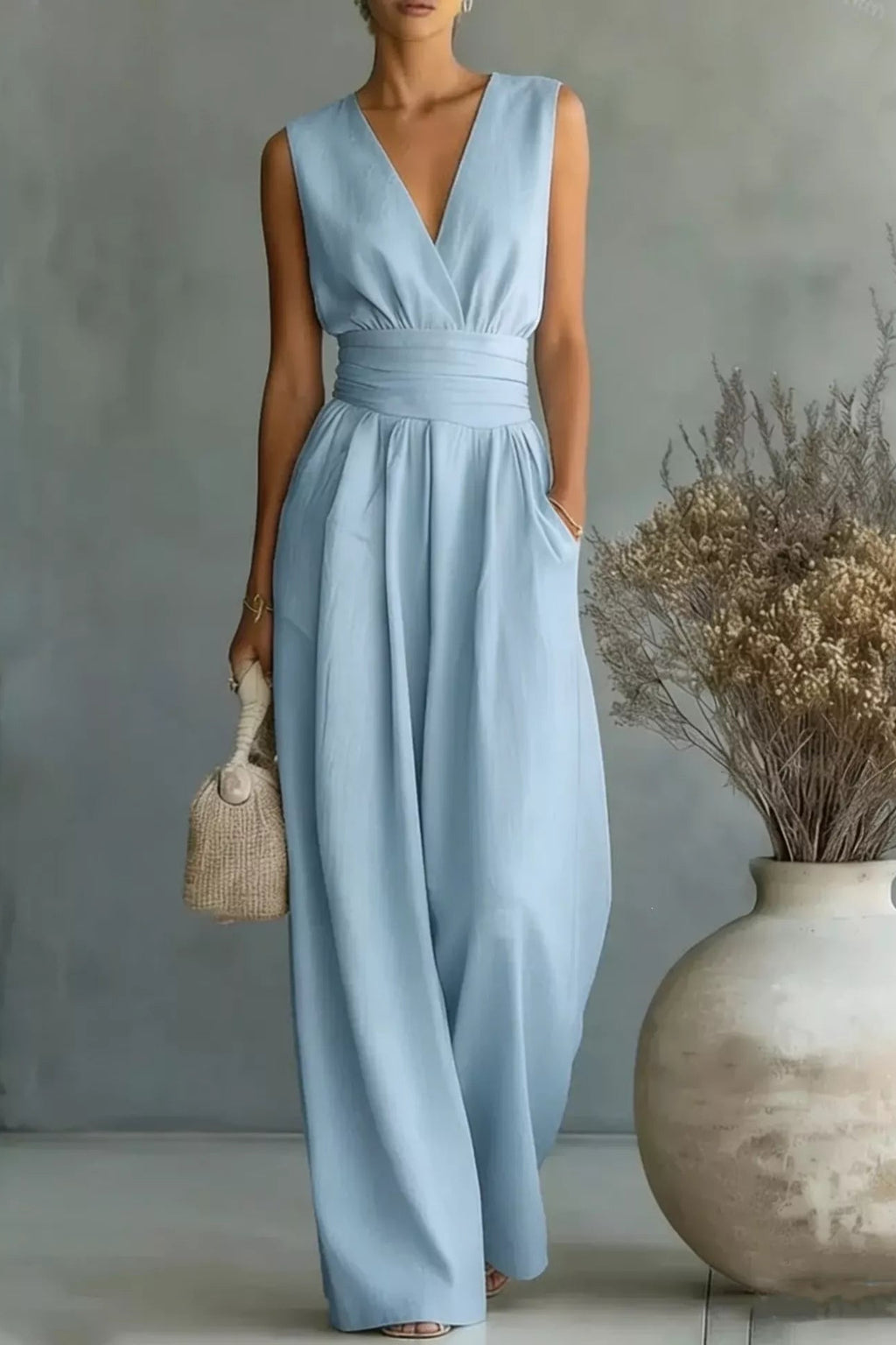 Woman wearing a light blue dress holding a woven bag in a minimalistic setting with a vase and dried plants.