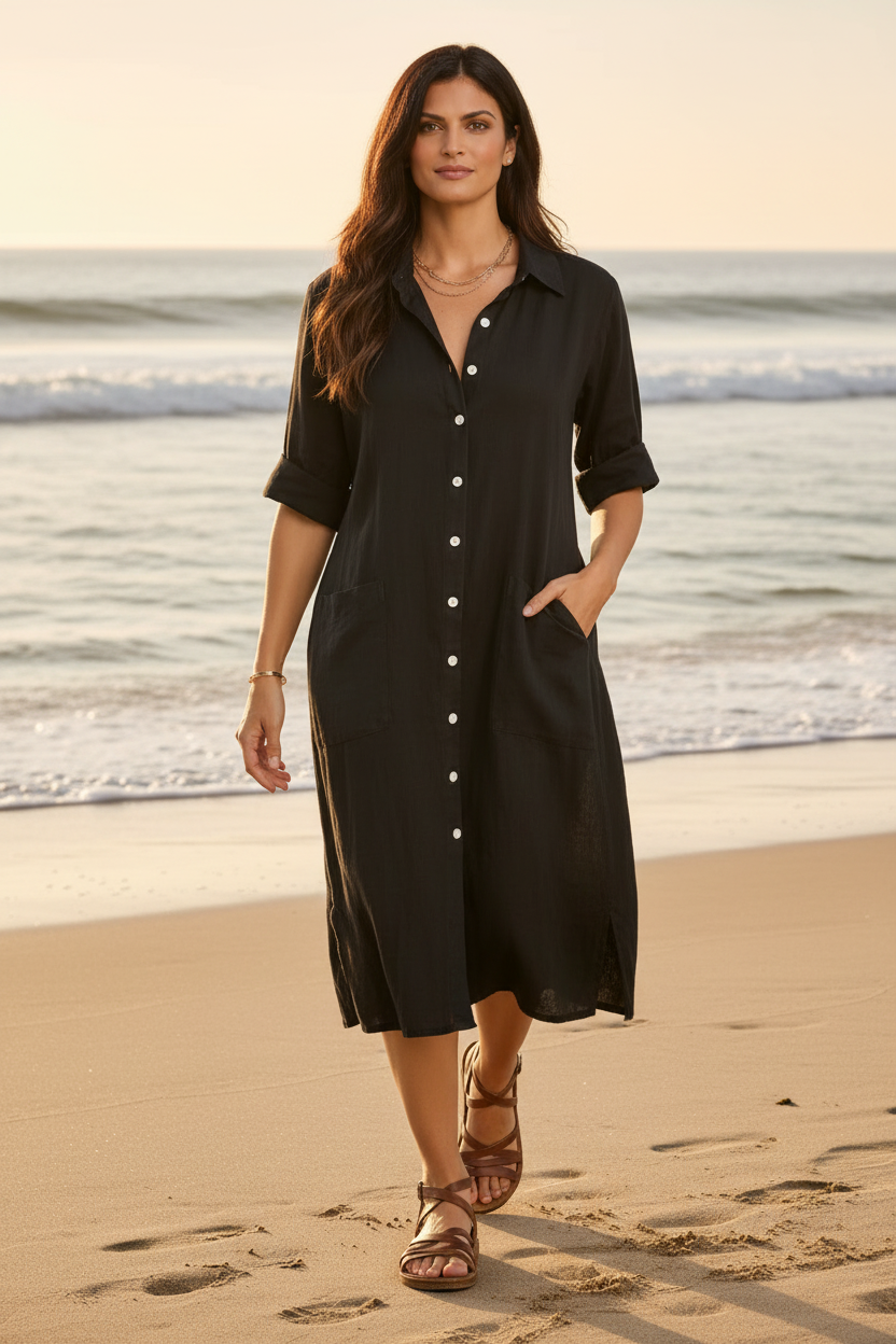 A woman stands on a sandy beach wearing a black button-front midi shirt dress with rolled sleeves and side pockets. The ocean and warm golden sunset light are visible in the background.