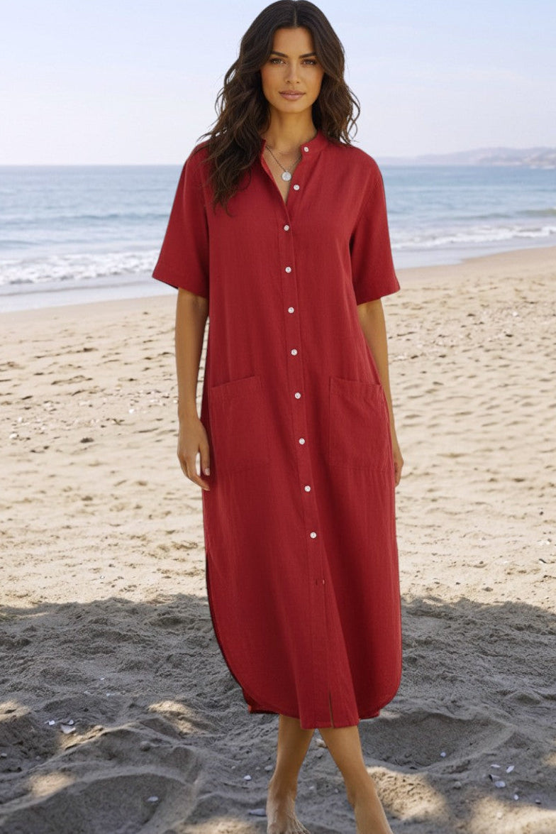 A woman wearing a red short-sleeve button-front midi dress stands barefoot on a sandy beach with the ocean in the background.