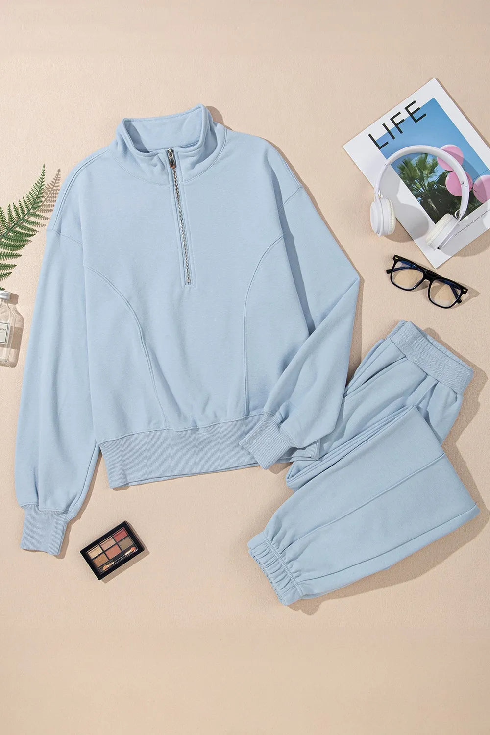 Light blue tracksuit with a magazine, glasses, and makeup palette on a beige background