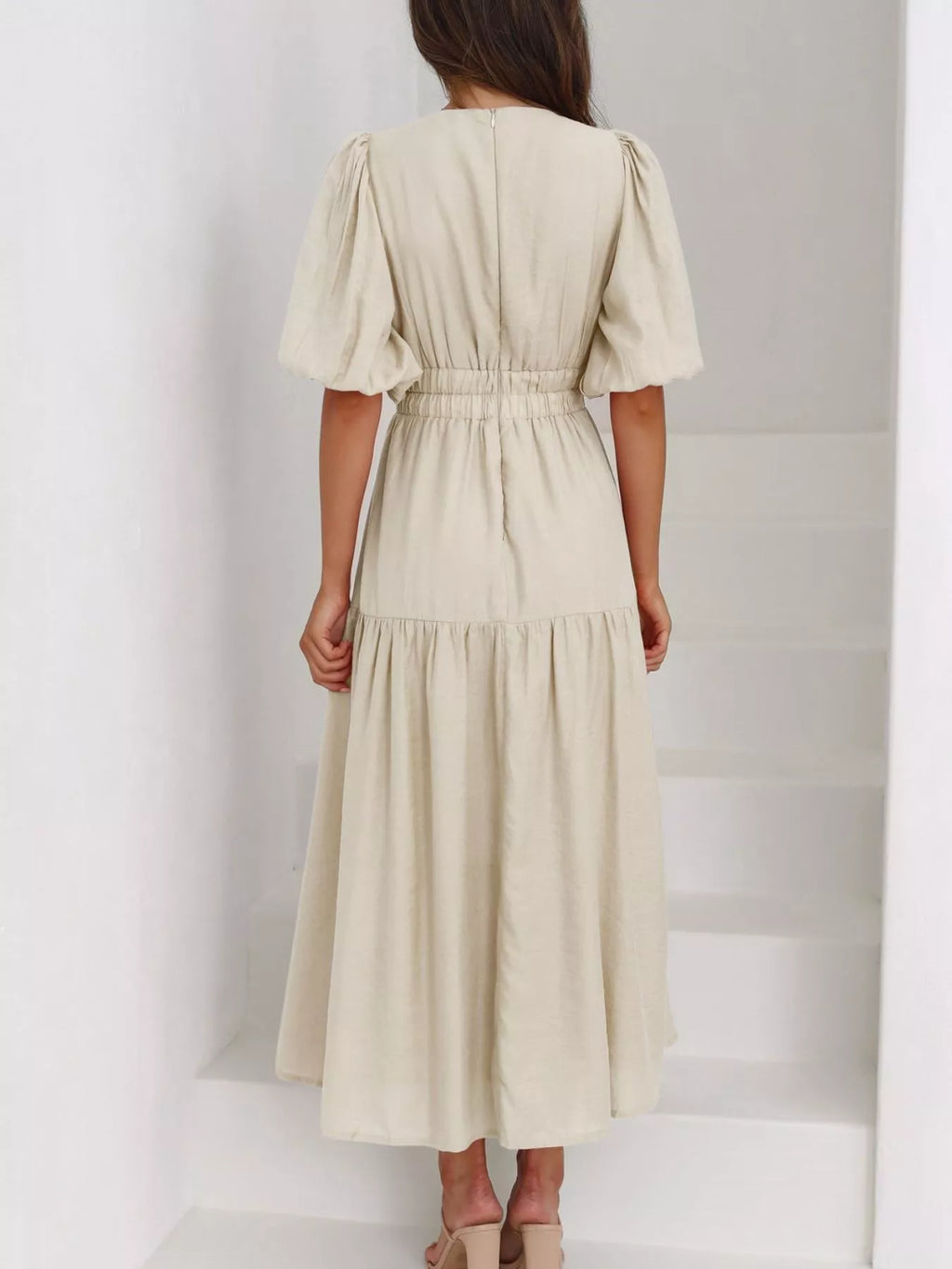 A woman viewed from behind wearing a light beige midi dress with short puff sleeves, a gathered waist, and a tiered skirt, standing on white stairs.