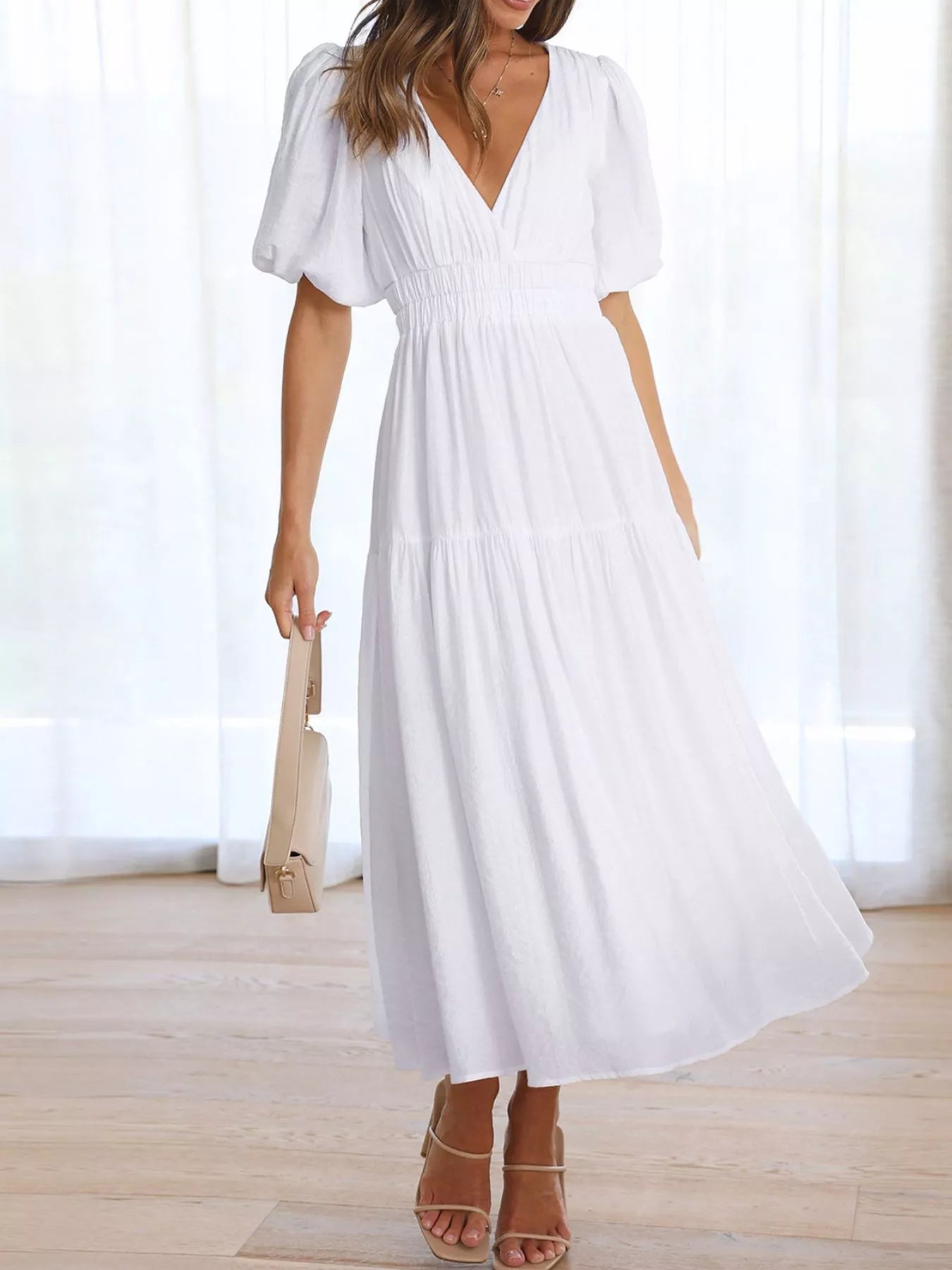 A woman wearing a white midi dress with puff sleeves, a deep V-neckline, and a smocked waist, holding a beige handbag in a bright room with light wood floors and sheer curtains.