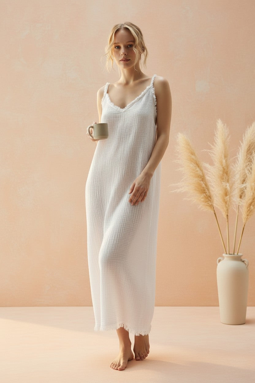 A woman stands barefoot in a studio wearing a white textured slip dress with thin straps and a frayed hem, holding a small mug. The background is a soft peach-pink wall with a beige vase of dried pampas grass on the right.