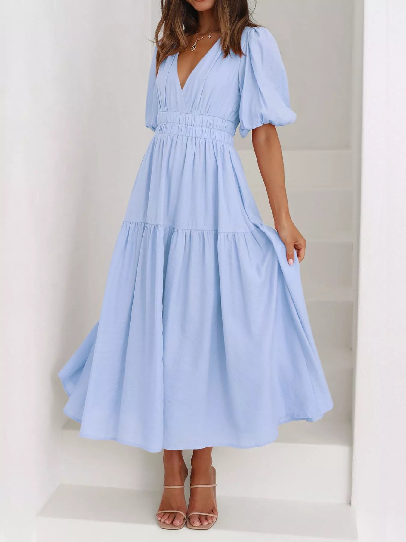A woman wearing a light blue tiered maxi dress with puff sleeves and a deep V-neckline, standing on white stairs.