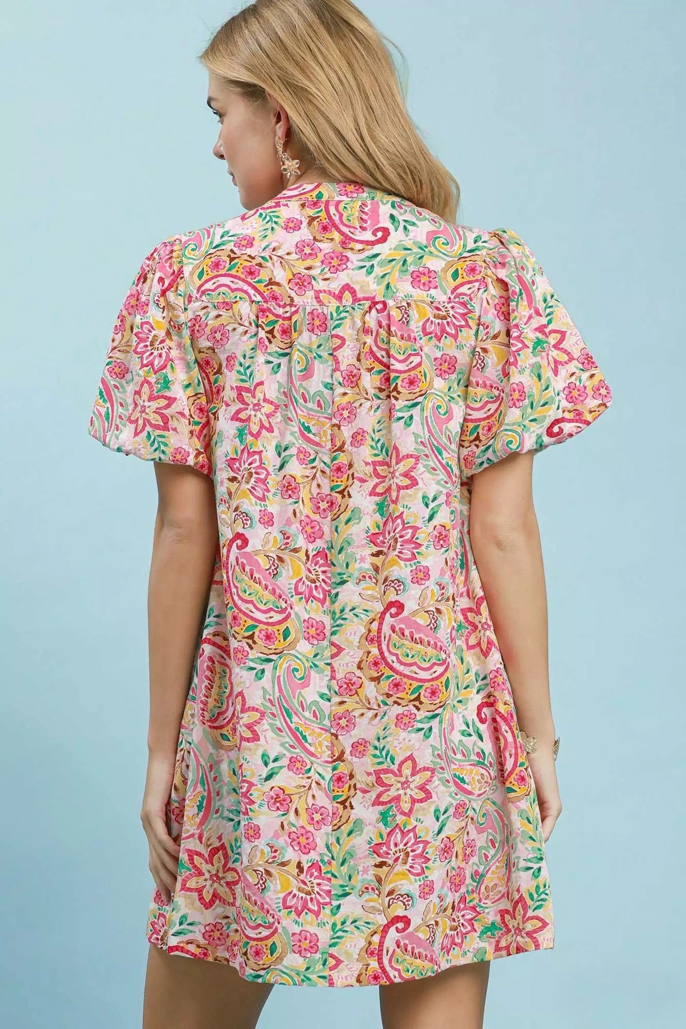 A woman shown from the back wearing a short-sleeve midi dress with a colorful paisley and floral print on a light cream background, set against a solid light blue backdrop.