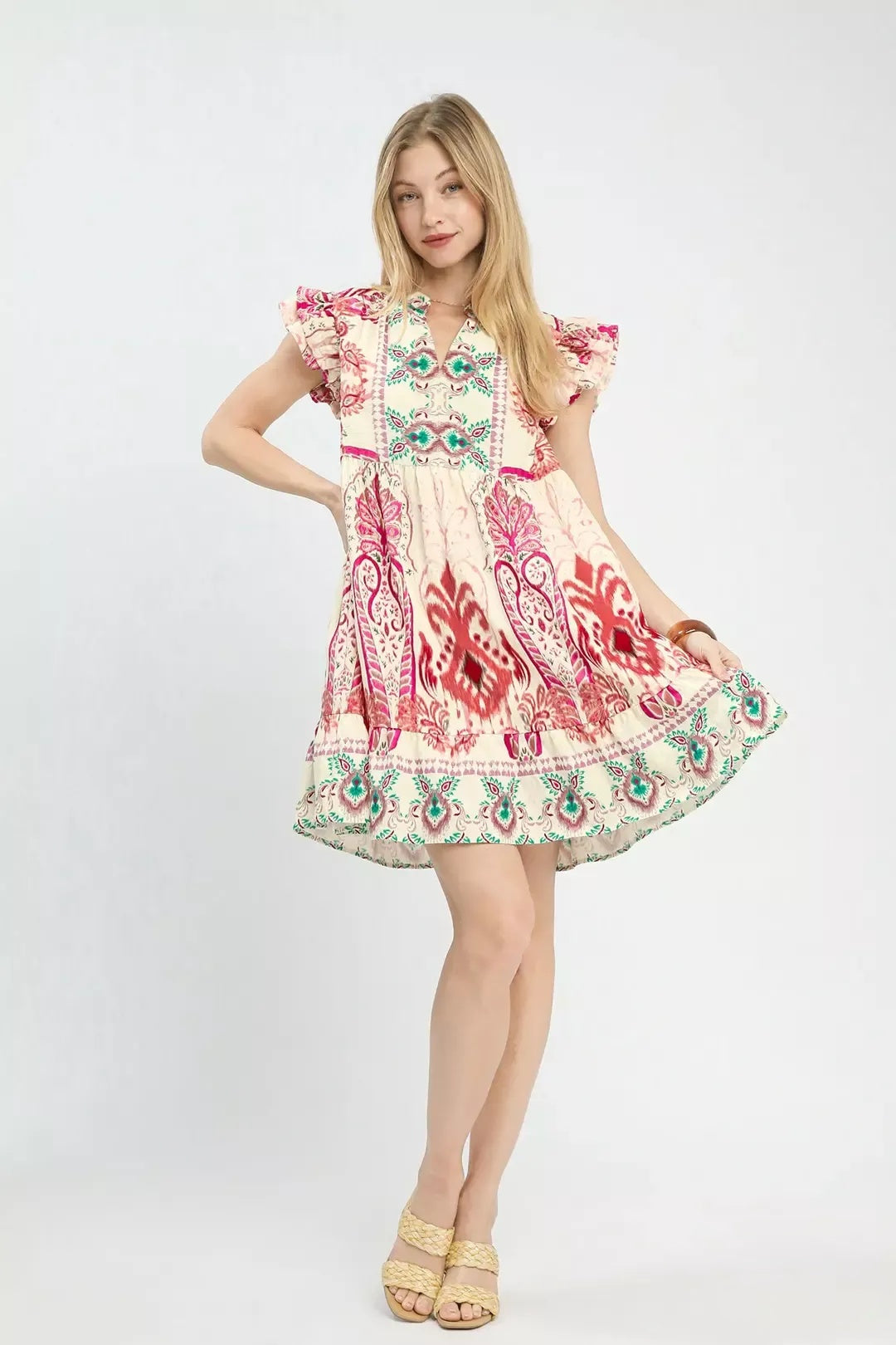 A young girl modeling a short, flutter-sleeve dress with a bohemian-style multicolor print in red, green, and blue on a cream background, paired with gold sandals, against a light gray studio backdrop.