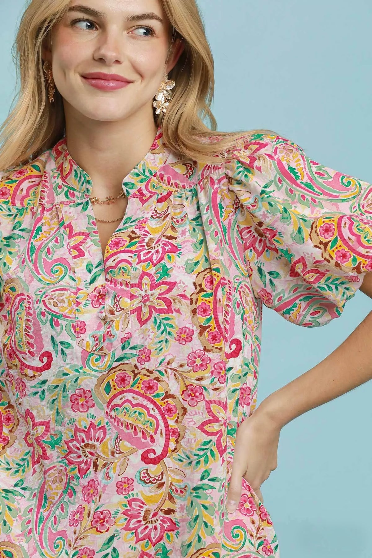 A woman wearing a short-sleeve button-front blouse with a colorful paisley and floral print on a light pink base, posed against a solid light blue background.