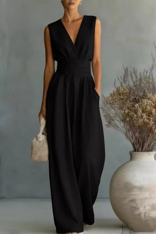 Woman wearing a black sleeveless jumpsuit holding a beige handbag against a neutral background.