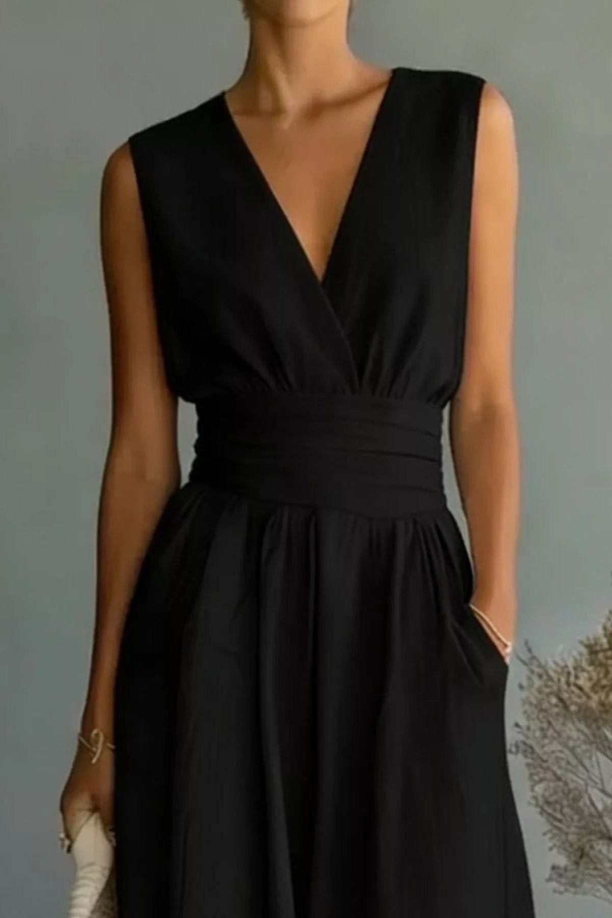 Woman wearing a black sleeveless jumpsuit holding a beige handbag against a neutral background.