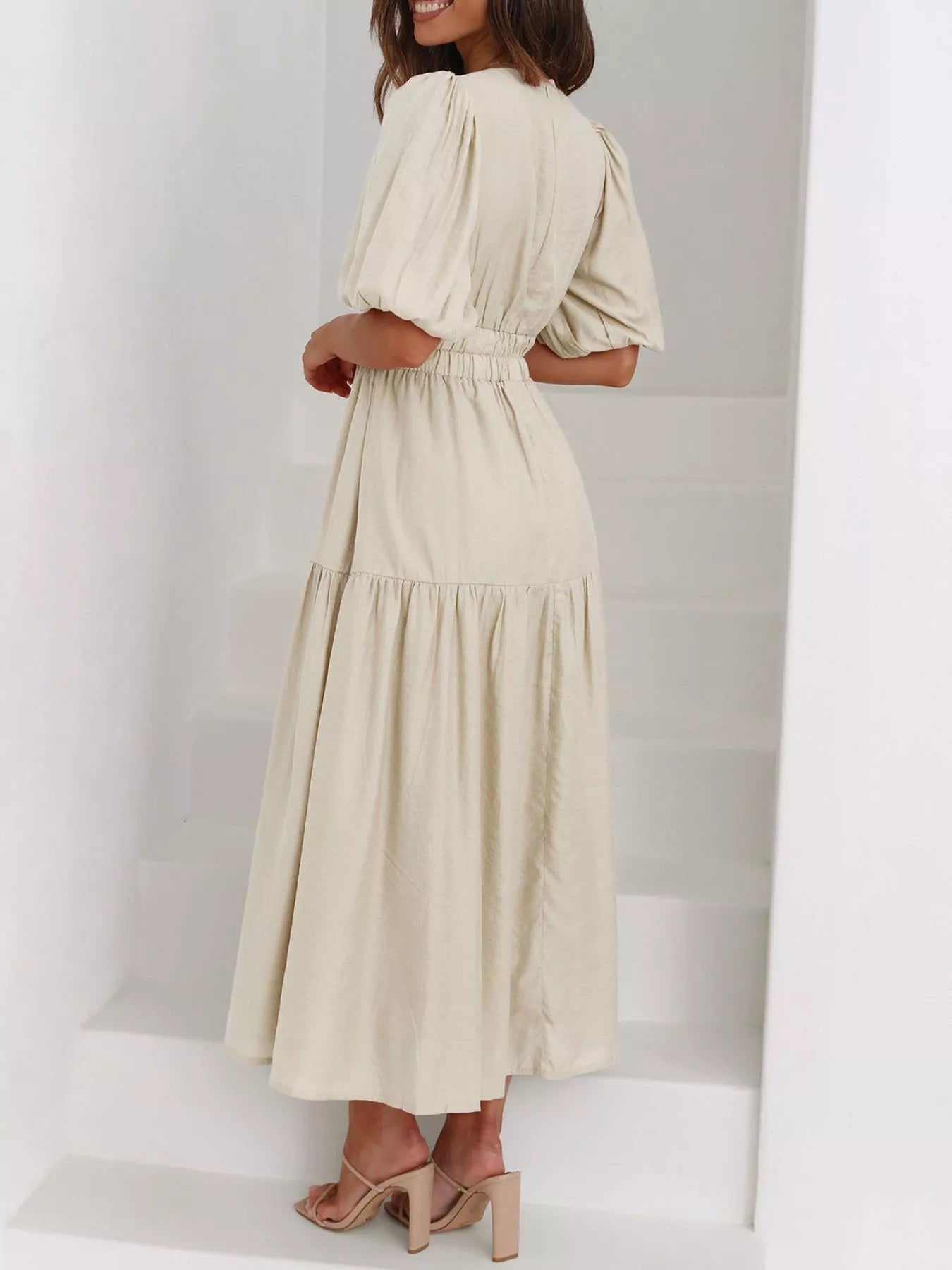 A woman wearing a light beige midi dress with puff sleeves and a tiered skirt, standing on white stairs.