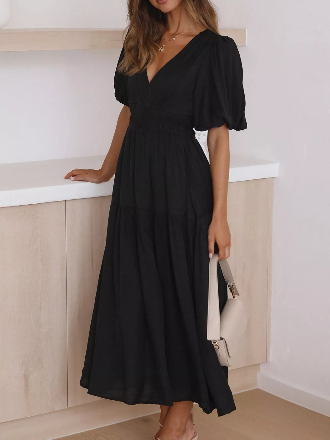 A woman wearing a black midi dress with puff sleeves and a V-neckline, standing indoors near a light wood counter and holding a small light-colored handbag.