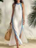 A woman wearing a sleeveless white maxi dress with light blue trim, standing on a sandy surface with palm leaves in the background and holding a woven straw bag.