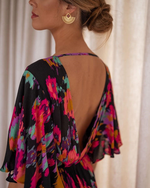 A woman shown from the back wearing a black dress with a vibrant, multicolored floral print in pink, purple, teal, and orange. She is wearing gold huggie hoop earrings and standing in front of a soft beige curtain background.