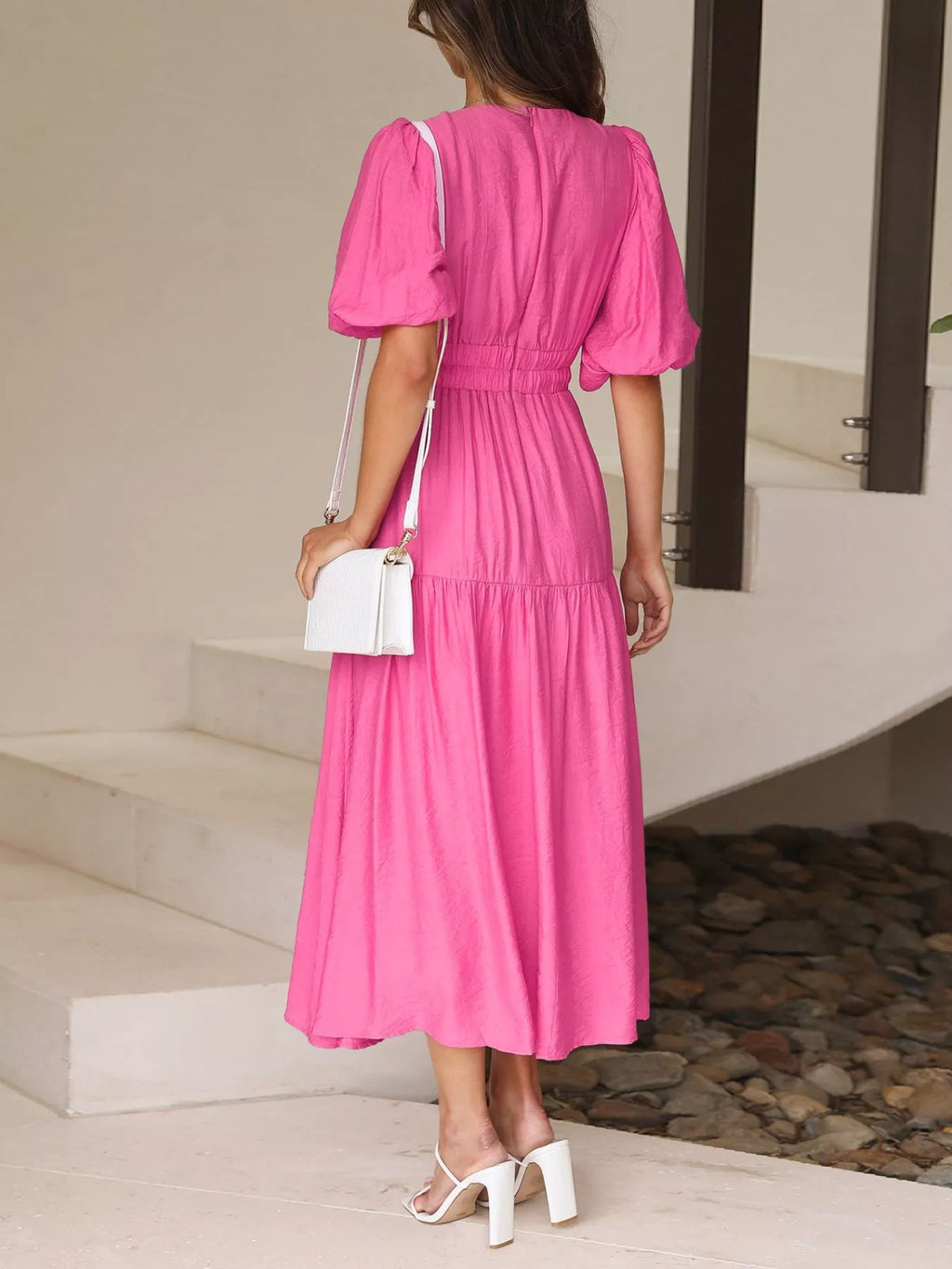 A woman wearing a bright pink midi dress with puff sleeves and a gathered waist, shown from the back in an outdoor setting with white high heels and a white handbag.