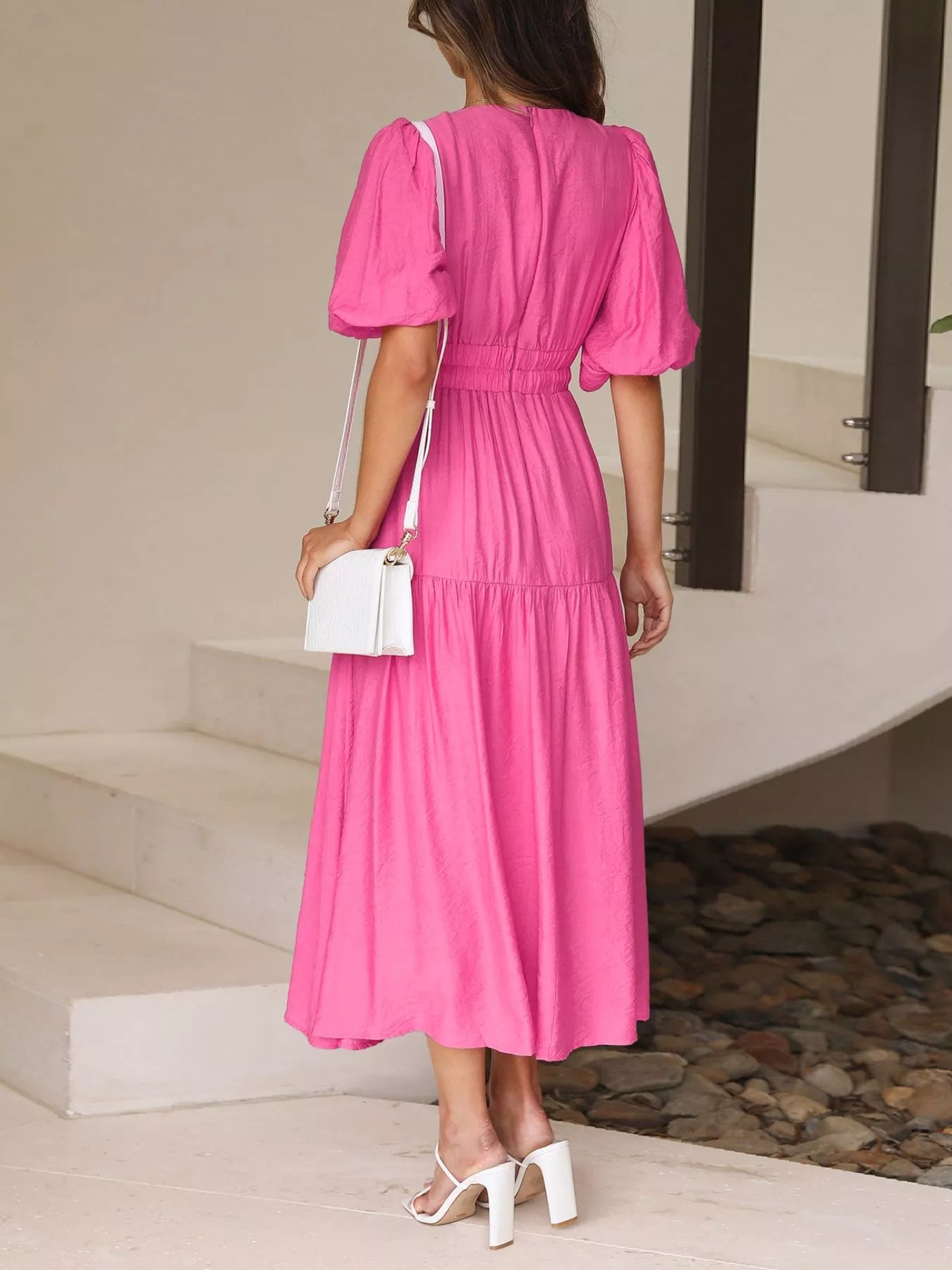 A woman wearing a bright pink midi dress with puff sleeves and a gathered waist, shown from the back in an outdoor setting with white high heels and a white handbag.