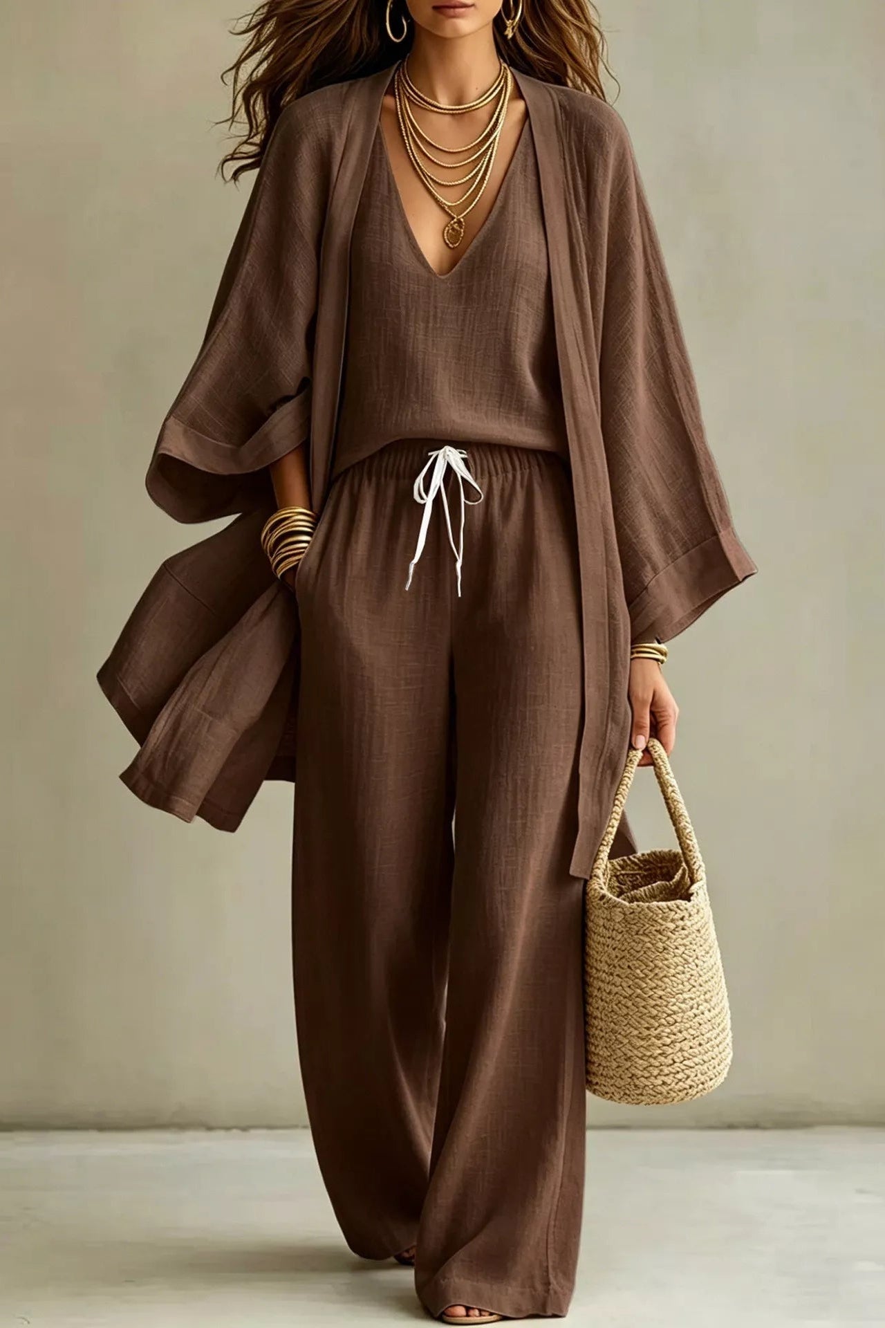 Woman wearing a brown three-piece set with open-front kimono cardigan, V-neck cami, and wide-leg drawstring pants, holding a woven tote bag