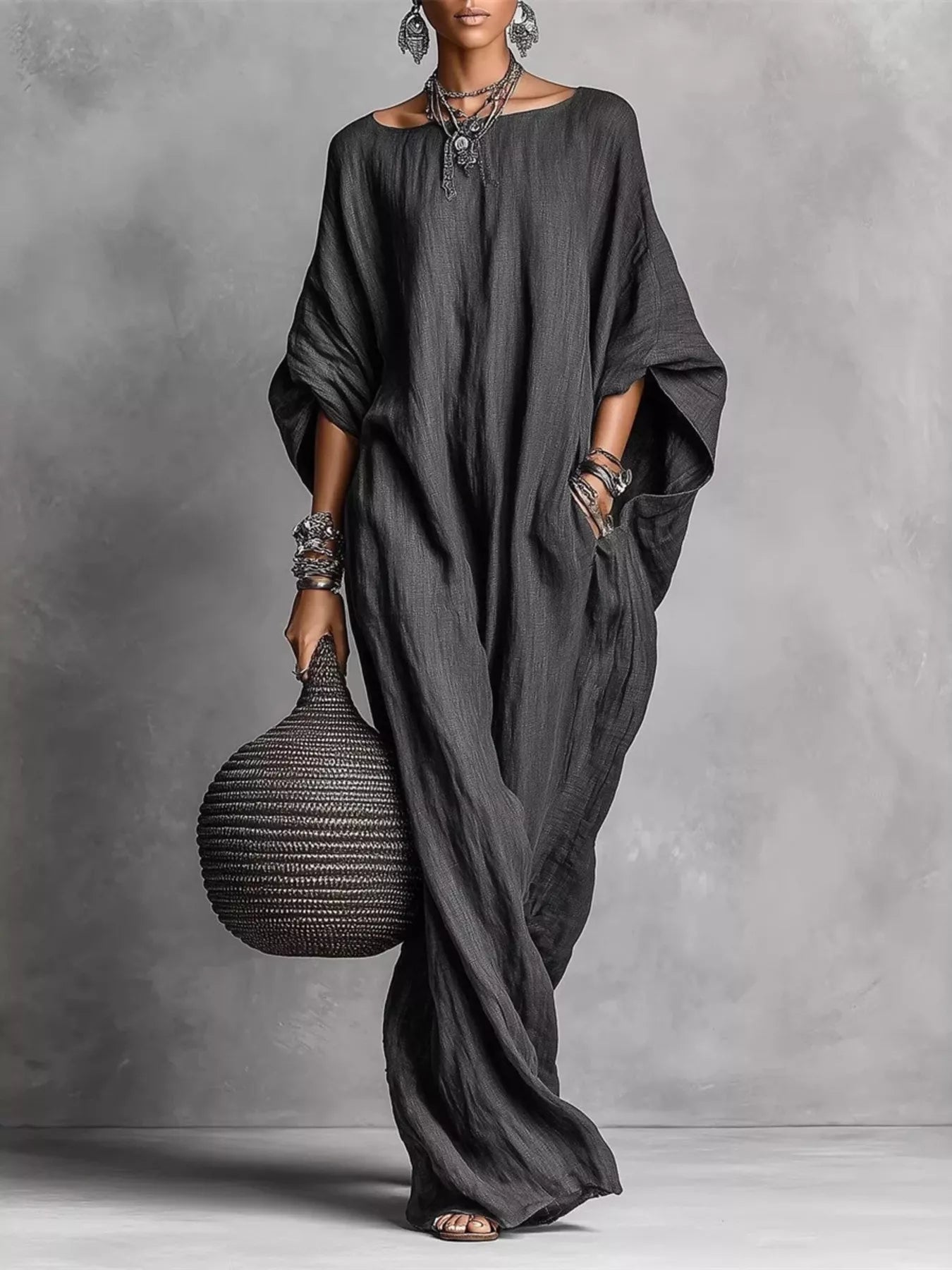A woman models a dark charcoal gray, loose-fitting linen caftan dress with wide draped sleeves, holding a woven dark brown basket bag against a textured gray studio backdrop.