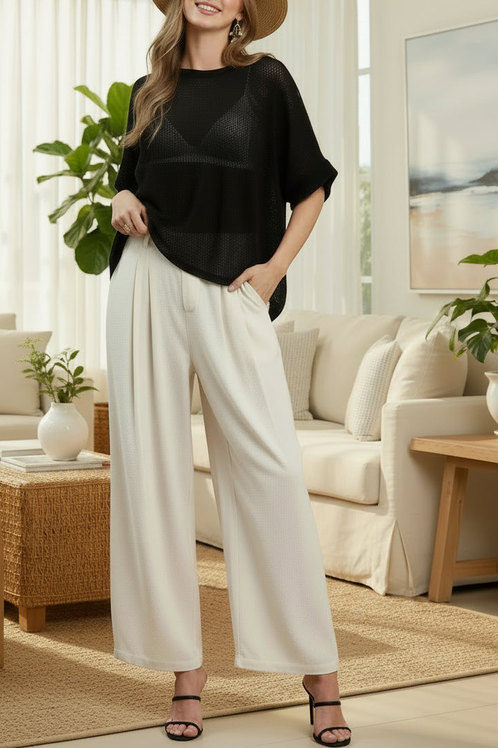 A woman modeling a black short-sleeve top and cream wide-leg pants against a solid warm brown studio background, wearing a straw hat and brown sandals. The word 'OATMEAL' appears in the bottom right corner.
