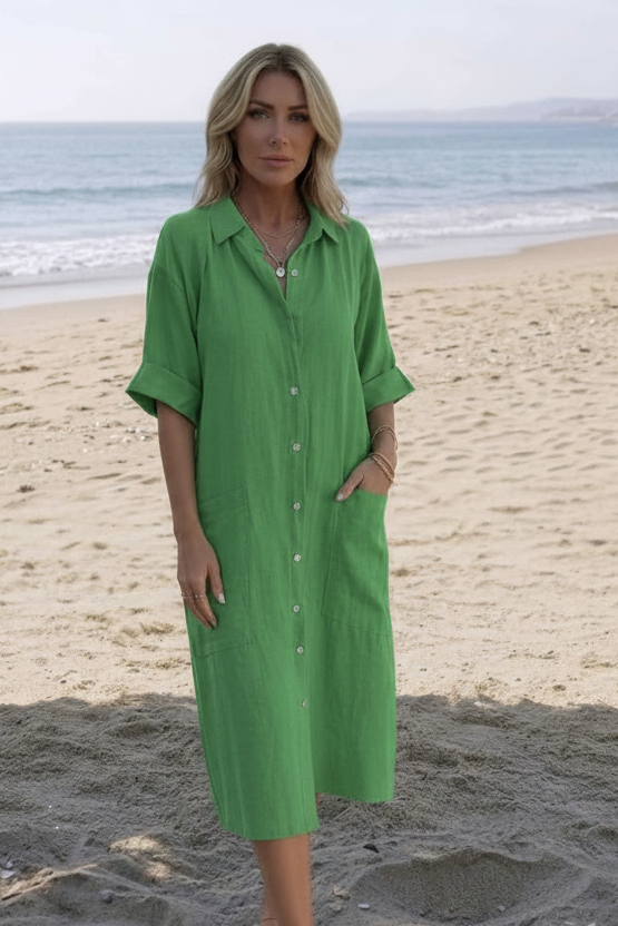 A woman wearing a green linen button-down shirt dress stands on a sandy beach with the ocean in the background.