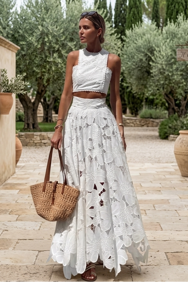 A woman stands in a sunlit stone courtyard wearing a white sleeveless crop top and a long white embroidered skirt, holding a woven straw tote bag. Olive trees, beige walls, and large terracotta pots are visible in the background.