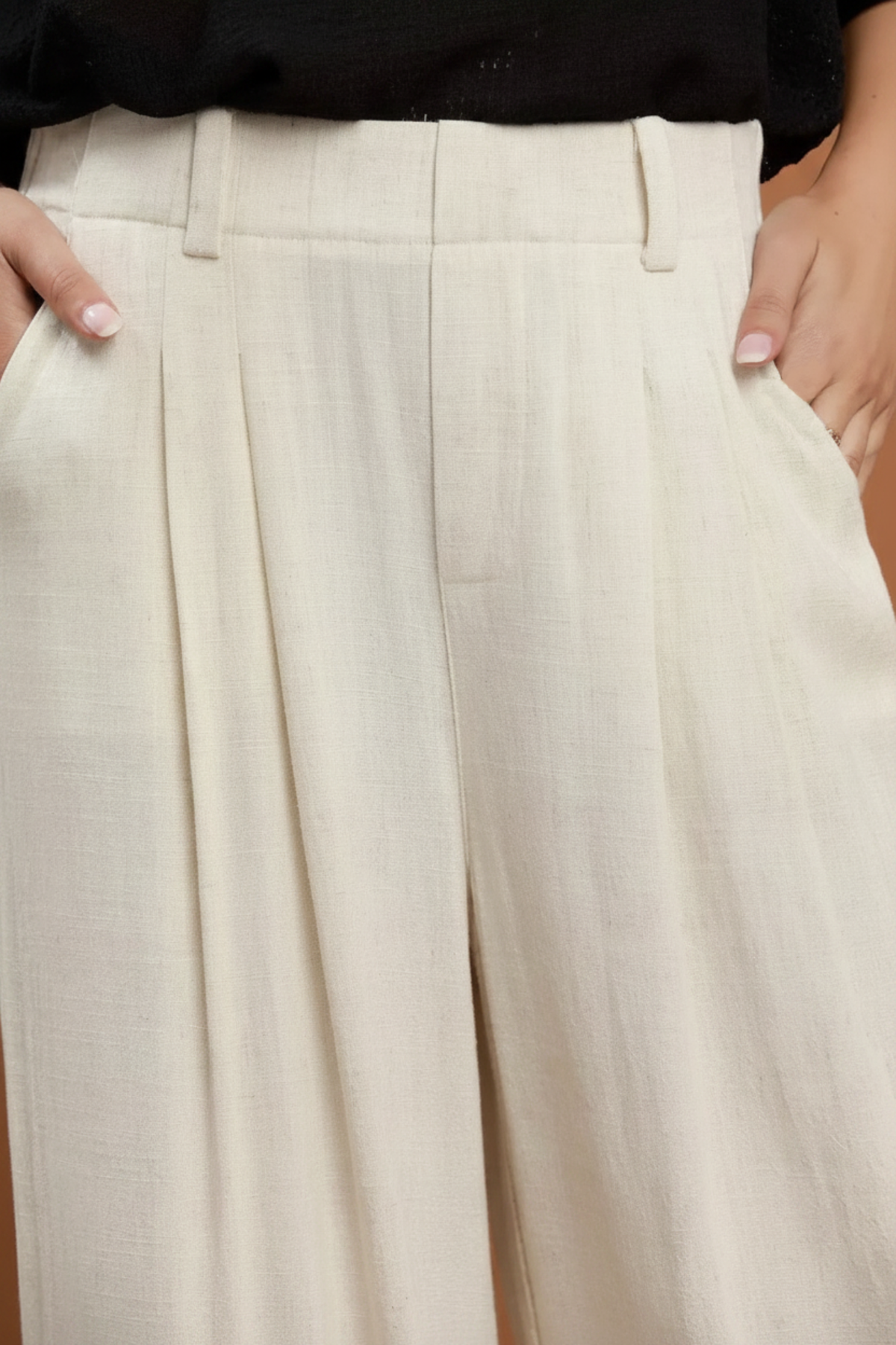 Close-up of a person wearing high-waisted, wide-leg pleated trousers in a light cream or off-white fabric, paired with a black top.