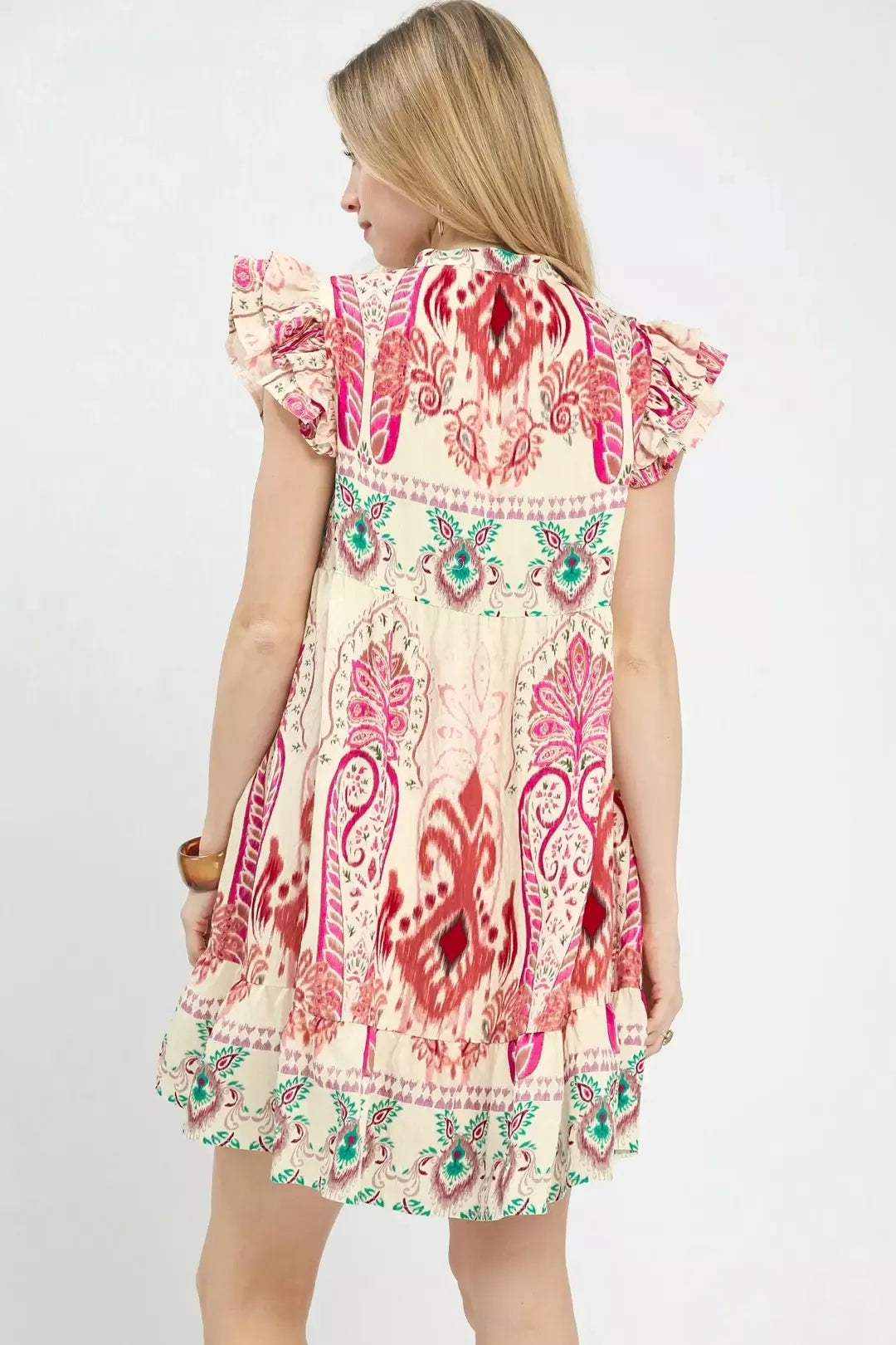 A woman shown from the back wearing a short-sleeve cream boho dress with a vibrant pink, red, and green paisley pattern and ruffled trim.
