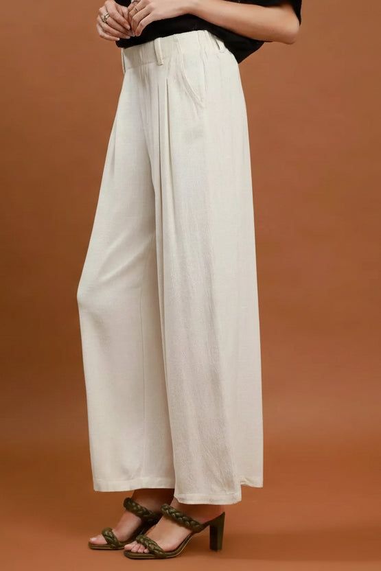 A person wearing high-waisted, wide-leg ivory linen pants with a black top and green heeled sandals, standing against a solid terracotta background.